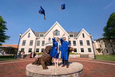 Two graduates in blue gowns tossing their grad caps in the air at the Pride lion statues