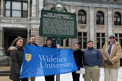 Students holding a blue Widener University flag pose in front of a New Hampshire sign