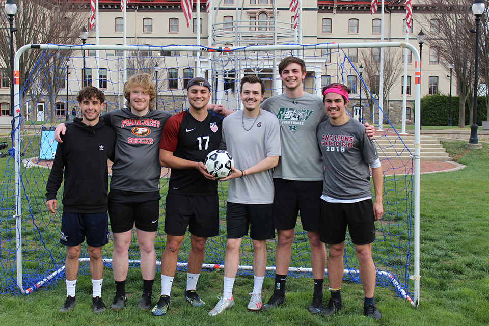 Six students pose for a photo in a soccer net, holding a soccer ball