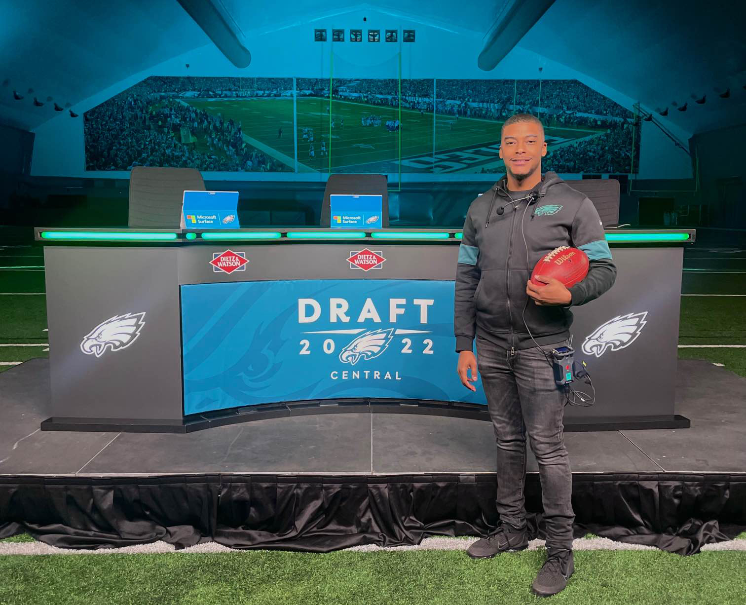 Alum Andrew Proctor holds a football and wears an Eagles jacket while standing in front of a desk with a sign for the NFL draft