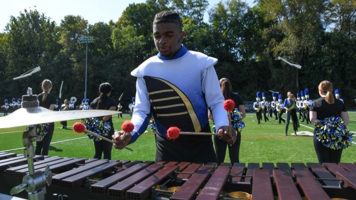 Band member plays xylophone while color guard, cheerleaders, and other band members perform at halftime behind