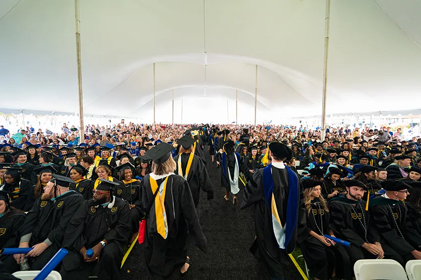 Back of faculty members walking down the center aisle past graduates and guests seated beneath a tent