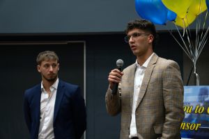 A student wearing a tan suit jacket speaks into a microphone while another student stands beside him