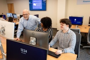 Two students sit in front of a computer in Widener University's finance lab, as a professor stands beside them, pointing at the screen.