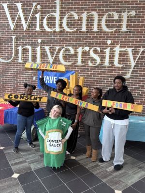 Six Widener University students pose beneath the university's sign, holding colorful signs highlighting consent principles. One student wears a green "Consent Sauce" costume, while others hold signs reading "Consent," "Reversible," "Freely Given," and "Informed."