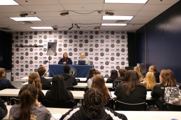 Students face a table on a stage where presenters are speaking