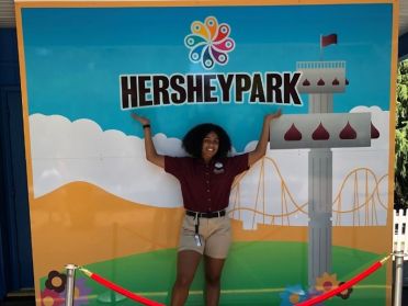 Donya Moore stands in front of a Hershey Park sign with her hands in the air