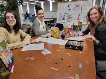 Three students sitting at a library table with puzzles and coloring supplies on it.