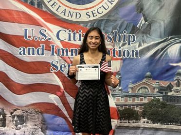 Student holds a citizenship document while standing in front of a background with American flag and other Americana