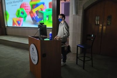 Masked male student standing at podium in Lathem Hall with bright screen of his presentation
