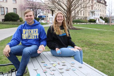 Two students sit atop a table holding painted rocks, with other painted rocks displayed before them