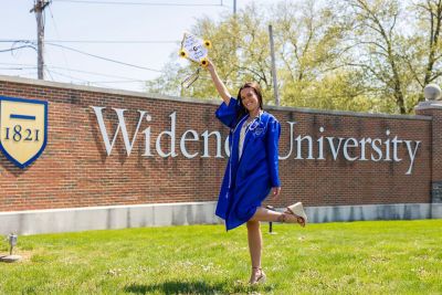 Kelsey Byrd poses in front of the Widener University sign in cap, gown and stethoscope.