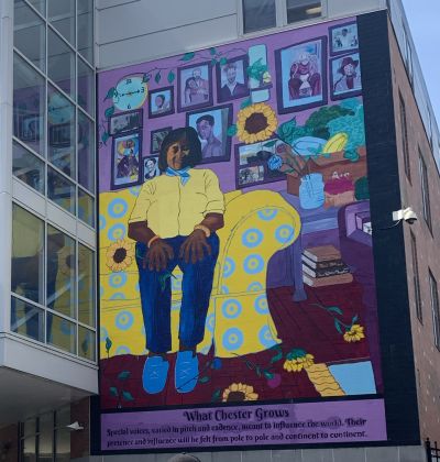 Mural on the side of Metro Hall featuring a woman seated on a yellow couch in a room, with portraits behind her and flowers and produce around her.