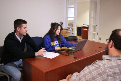 Students sit across from a client at a dark wooden desk. There are papers and a laptop on the desk