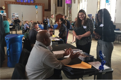 Two students in professinal attire approach a table and talk to a prospective employer