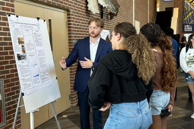 A student presents their research to two student audience members in front of a research poster.