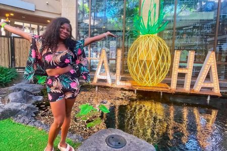 Kendall Gordon stands next to a small pond of water and an 'aloha' sign during a study abroad trip