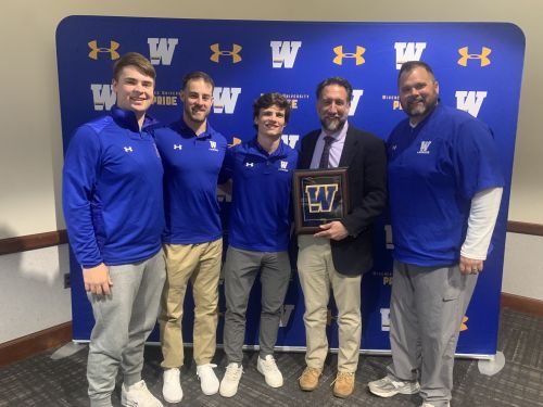 Vice Provost Mark Nicosia and members of the men's lacrosse team pose for a photo in front of a Widener Athletics backdrop