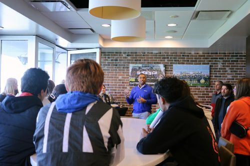 Students sit around a table listening to a professor talking