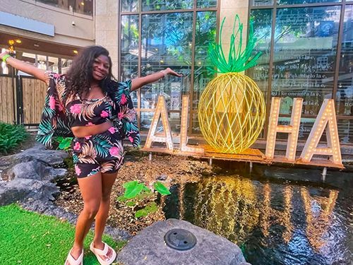 Kendall Gordon stands next to a small pond in front of a sign that says ‘aloha’ on a study abroad trip
